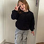 child, peace_sign, smiling, indoor, door, black_sweater, gray_pants, shoes, floor_tiles, furry_stool, happy, person, casual_clothing, standing, pointing, young_child, long_hair, light_hair, home_interior, gesture