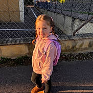 Joy participe au concours pour gagner de l'argent avec cette photo : child, girl, braids, pink_hoodie, pink_backpack, brown_boots, chain_link_fence, sidewalk, sunlight, shadow, outdoor, urban, casual_clothing, standing, portrait, daytime, smile, young, cute, street