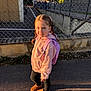 child, girl, braids, pink_hoodie, pink_backpack, brown_boots, chain_link_fence, sidewalk, sunlight, shadow, outdoor, urban, casual_clothing, standing, portrait, daytime, smile, young, cute, street