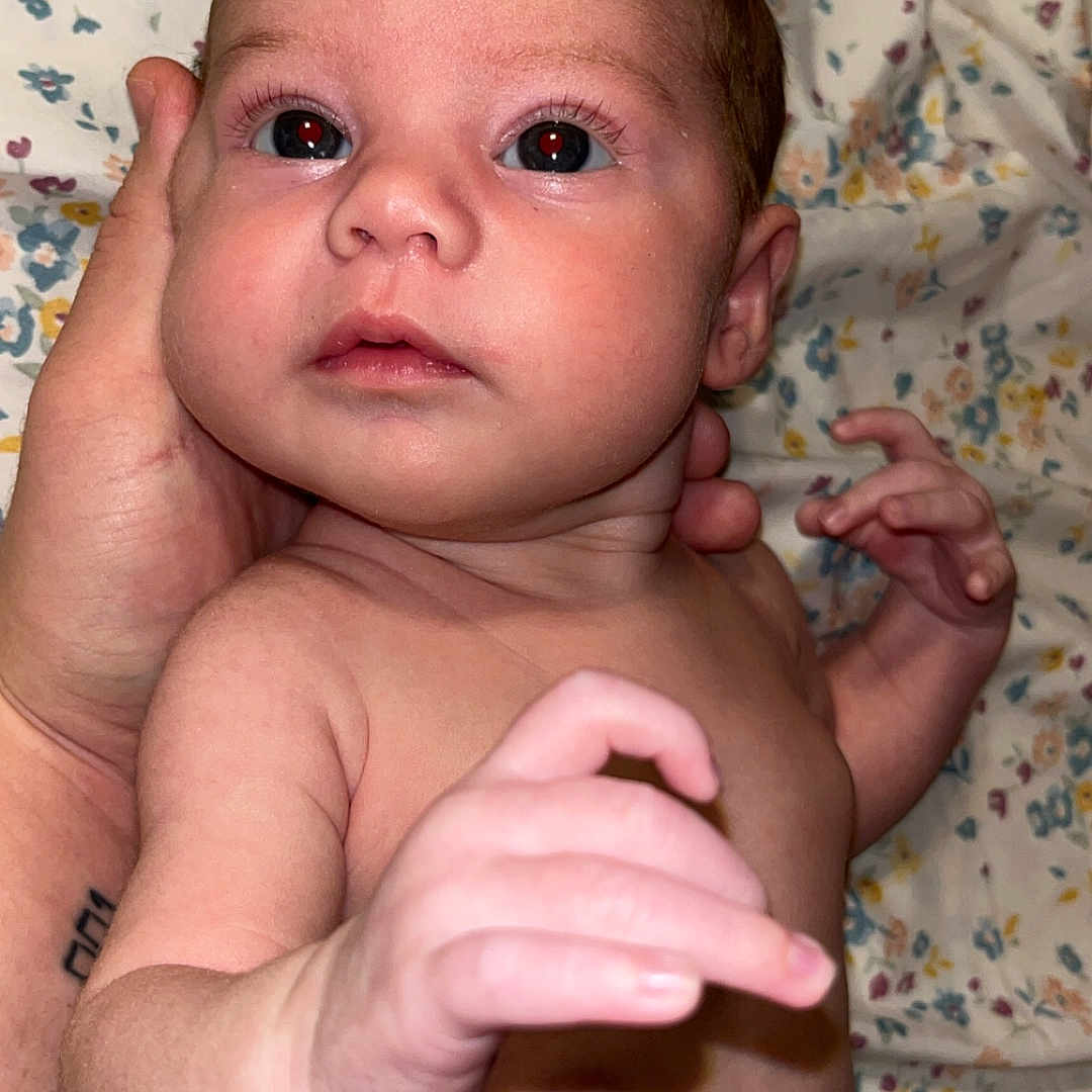 Brya is registered to the contest to win money with this photo: adult_hand, baby, blanket, child, closeup, cute, diaper, expression, eyes, face, floral_pattern, hand, human, indoor, infant, lying_down, newborn, portrait, skin, skin_texture