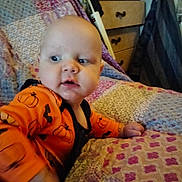 Waylon is registered to the contest to win money with this photo: baby, infant, child, orange_clothing, pumpkin_print, quilt, bedspread, furniture, drawer, curious, indoor, person, portrait, head, face, hand, bedroom, cozy, patterned, home