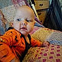 baby, infant, child, orange_clothing, pumpkin_print, quilt, bedspread, furniture, drawer, curious, indoor, person, portrait, head, face, hand, bedroom, cozy, patterned, home