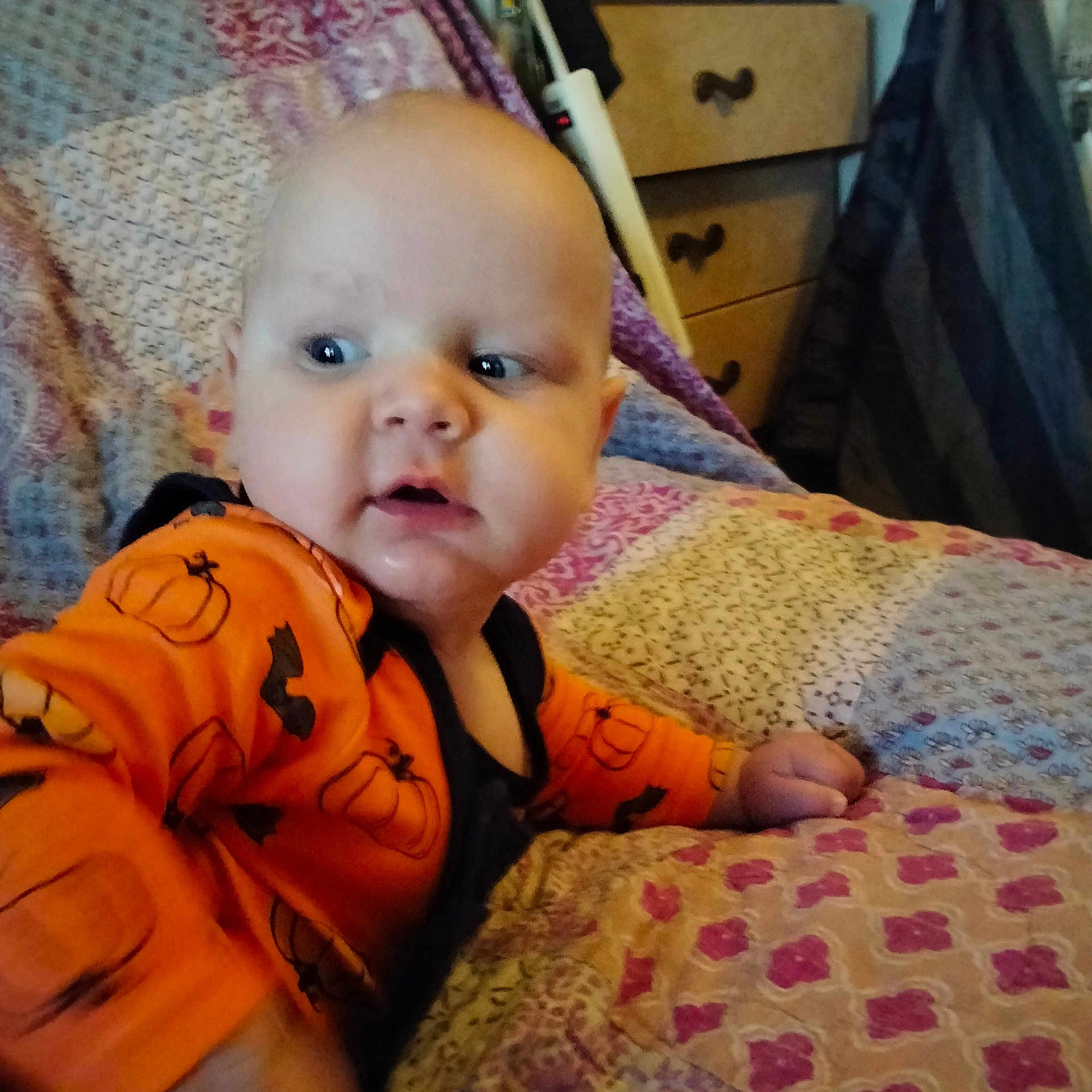Waylon is registered to the contest to win money with this photo: baby, infant, child, orange_clothing, pumpkin_print, quilt, bedspread, furniture, drawer, curious, indoor, person, portrait, head, face, hand, bedroom, cozy, patterned, home