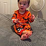 barefoot, bat_pattern, bed, bedroom, child, curly_hair, cute, door, expression, fan, gray_bedspread, halloween, holding_object, indoor, light_skin, orange_clothing, pajamas, sitting, toddler, young_child