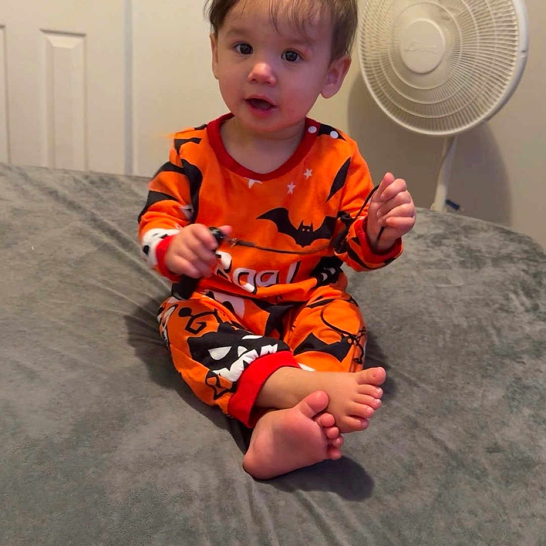 Madeleine joined the competition — help win amazing prizes! barefoot, bat_pattern, bed, bedroom, child, curly_hair, cute, door, expression, fan, gray_bedspread, halloween, holding_object, indoor, light_skin, orange_clothing, pajamas, sitting, toddler, young_child