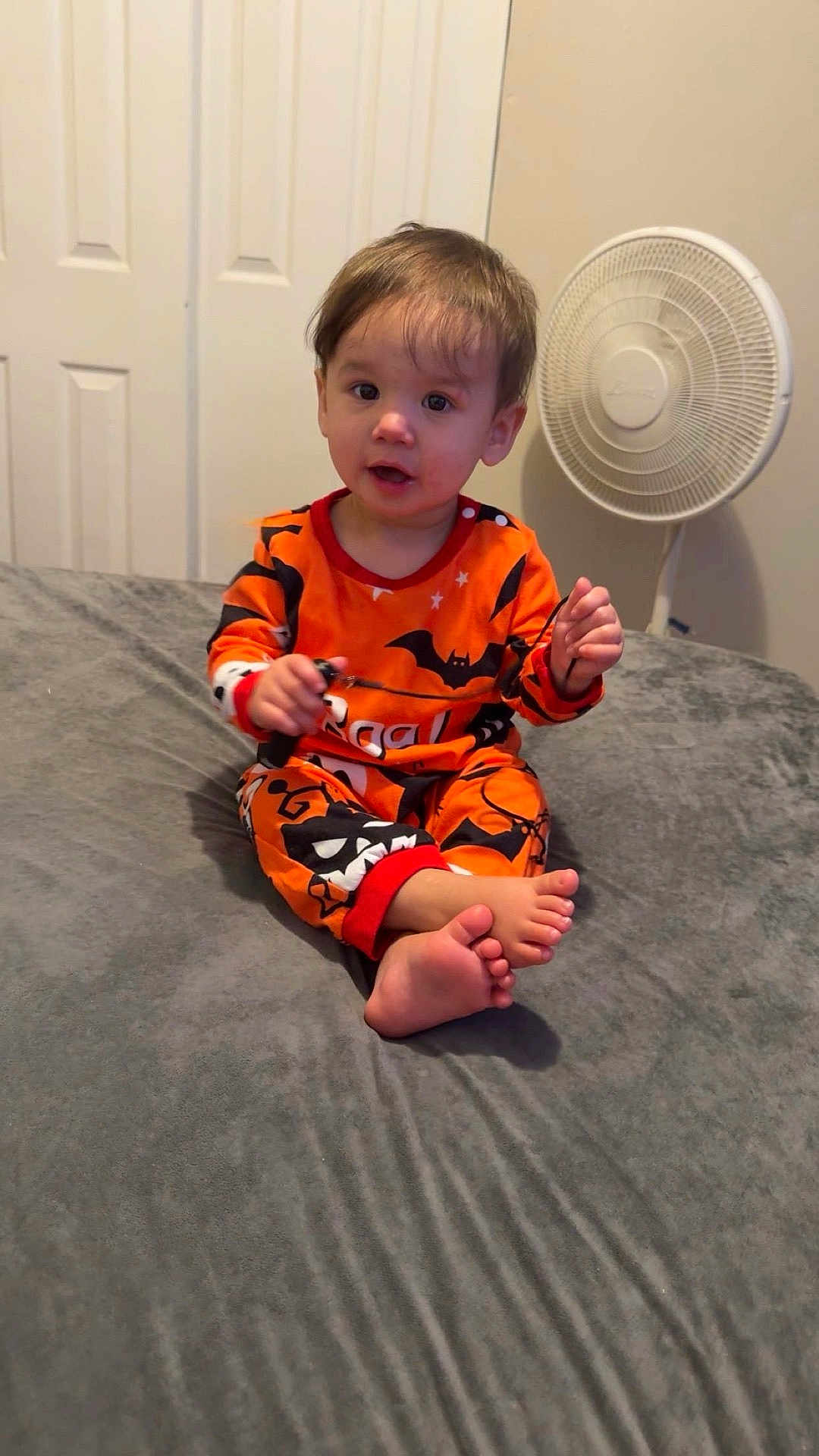 Madeleine joined the competition — help win amazing prizes! barefoot, bat_pattern, bed, bedroom, child, curly_hair, cute, door, expression, fan, gray_bedspread, halloween, holding_object, indoor, light_skin, orange_clothing, pajamas, sitting, toddler, young_child