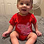 baby, child, red_clothing, onesie, peppa_pig, smiling, sitting, bed, gray_bedspread, indoor, fan, door, happy, person, infant, cute, short_hair, skin, feet, hand