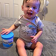 Madeleine joined the competition — help win amazing prizes! baby, child, smiling, onesie, sitting, indoor, fan, container, desitin, clothing, feet, hands, face, happy, room, bed, gray, hair, skin, expression