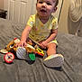 baby, child, yellow_dress, stuffed_animal, toy, bed, gray_bedspread, shoes, indoor, fan, door, person, smiling, sitting, cute, infant, playful, happy, home, young_child