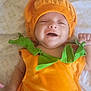 Hugo is registered to the contest to win money with this photo: baby, child, smiling, costume, pumpkin, orange, green, plush, bed, sheet, face, happy, infant, cute, lying_down, clothing, holiday, festive, person, indoors