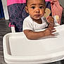 baby, high_chair, food, indoor, child, face, white_clothing, flooring, toy, pink, curly_hair, person, expression, hand, foot, table, seat, real_people, infant, eating