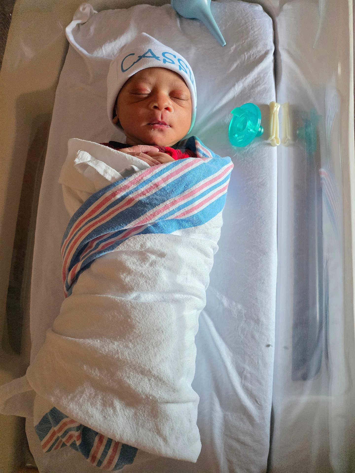 Cassius is registered to the contest to win money with this photo: newborn, baby, swaddled, blanket, hospital, bassinet, pacifier, sleeping, hat, infant, bed, resting, wrapped, cute, small, peaceful, person, child, indoors, healthcare