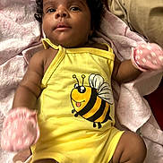 Malia is registered to the contest to win money with this photo: baby, infant, child, yellow_onesie, bee_graphic, pink_mittens, blanket, soft_fabric, cute, portrait, lying_down, indoor, person, skin, curly_hair, happy, closeup, smiling, comfortable, cozy