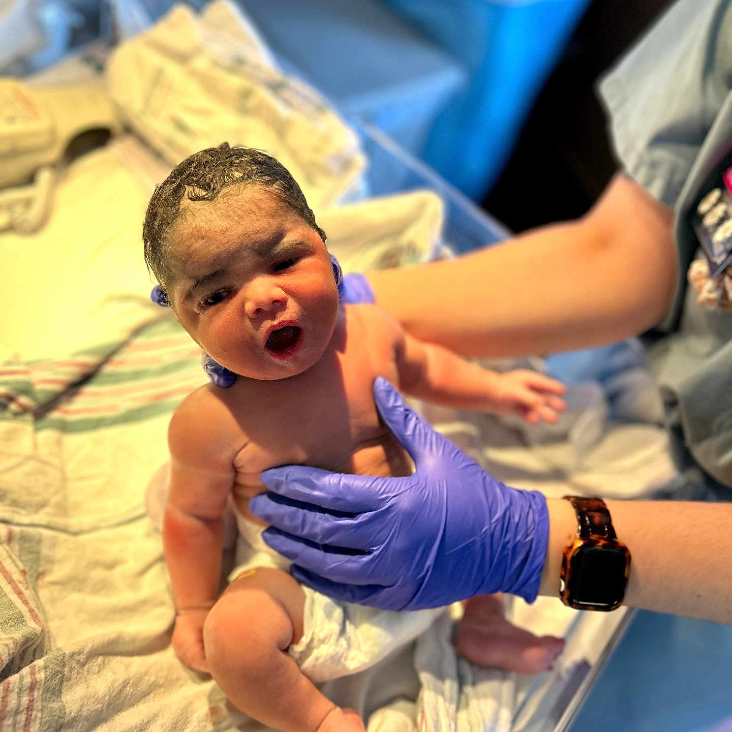 Malia is registered to the contest to win money with this photo: newborn, baby, hospital, medical_gloves, person, infant, bed, blanket, healthcare, care, human, skin, arm, hand, watch, diaper, expression, curious, indoor, nurse