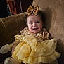 baby, child, smiling, yellow_dress, bow, sitting, armchair, happy, cute, indoors, clothing, face, portrait, infant, sock, furniture, headwear, toddler, expression, candid