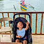 Lorenzo is registered to the contest to win money with this photo: toddler, stroller, child, river, boat, person, umbrella, wooden_railing, water, outdoor, nature, greenery, table, chair, baby_bib, footwear, daylight, smile, holding, relaxation