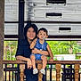 child, toddler, adult, pacifier, blue_clothing, wooden_railing, outdoor, greenery, smiling, person, balcony, daylight, hair, face, shoes, casual, holding, nature, portrait, relaxed