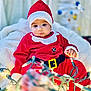 baby, blanket, child, christmas_lights, christmas_tree, cozy, cute, face, festive, headwear, holiday, holiday_decor, indoor, person, red_clothing, santa_claus_costume, seasonal, smiling, toddler, warm_lighting
