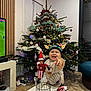 child, christmas_tree, decoration, elf_doll, elf_hat, festive, furniture, happy, holiday, indoor, lights, living_room, ornaments, pointing, smiling, table, television, toddler, toy, wooden_floor