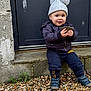 child, concrete, curious, door, hat, holding, jacket, outdoor, pants, pebbles, person, plants, pom_pom, rocks, shoes, sitting, smile, step, stone, toddler