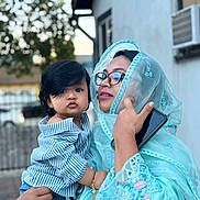 Mahbeer Rahman is registered to the contest to win money with this photo: accessory, blue_clothing, child, curly_hair, emotion, expression, face, family, glasses, hand, holding, outdoor, person, phone, portrait, striped_shirt, toddler, traditional_clothing, urban_background, woman