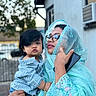 accessory, blue_clothing, child, curly_hair, emotion, expression, face, family, glasses, hand, holding, outdoor, person, phone, portrait, striped_shirt, toddler, traditional_clothing, urban_background, woman