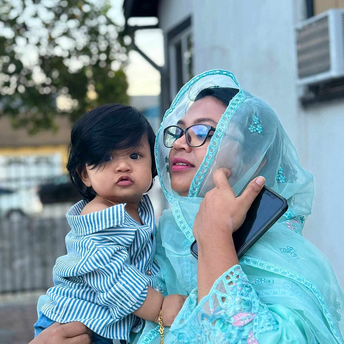 Mahbeer Rahman is registered to the contest to win money with this photo: accessory, blue_clothing, child, curly_hair, emotion, expression, face, family, glasses, hand, holding, outdoor, person, phone, portrait, striped_shirt, toddler, traditional_clothing, urban_background, woman