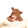 baby, infant, costume, bear, brown, cute, milestone, three_months, blanket, soft, chewing, hand, portrait, child, fuzzy, lying_down, curious, wooden_sign, studio_photo, white_background
