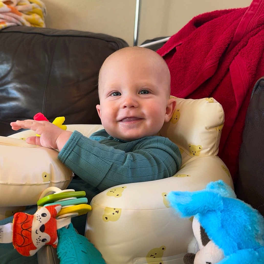 Jameson is registered to the contest to win money with this photo: baby, infant, smiling, toys, inflatable_seat, living_room, couch, lamp, red_blanket, blue_toy, yellow_duck_prints, child, happy, indoors, person, plush_toy, playtime, clothing, seated, cute