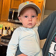 Jameson is registered to the contest to win money with this photo: baby, child, cap, onesie, kitchen, stove, cabinet, adult_hand, indoor, person, face, curious, holding, appliance, tile_floor, light, blue_eyes, clothing, home, portrait