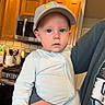 baby, child, cap, onesie, kitchen, stove, cabinet, adult_hand, indoor, person, face, curious, holding, appliance, tile_floor, light, blue_eyes, clothing, home, portrait