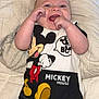 baby, infant, smiling, mickey_mouse, shirt, clothing, blanket, quilt, happy, face, hands, lying_down, cute, child, young, person, apparel, cartoon_character, black_pants, indoor