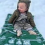 baby, child, snow, green_blanket, jacket, hood, ears, smile, happy, outdoor, winter_clothing, camouflage_overalls, socks, sitting, blanket, cold_weather, cute, infant, person, nature