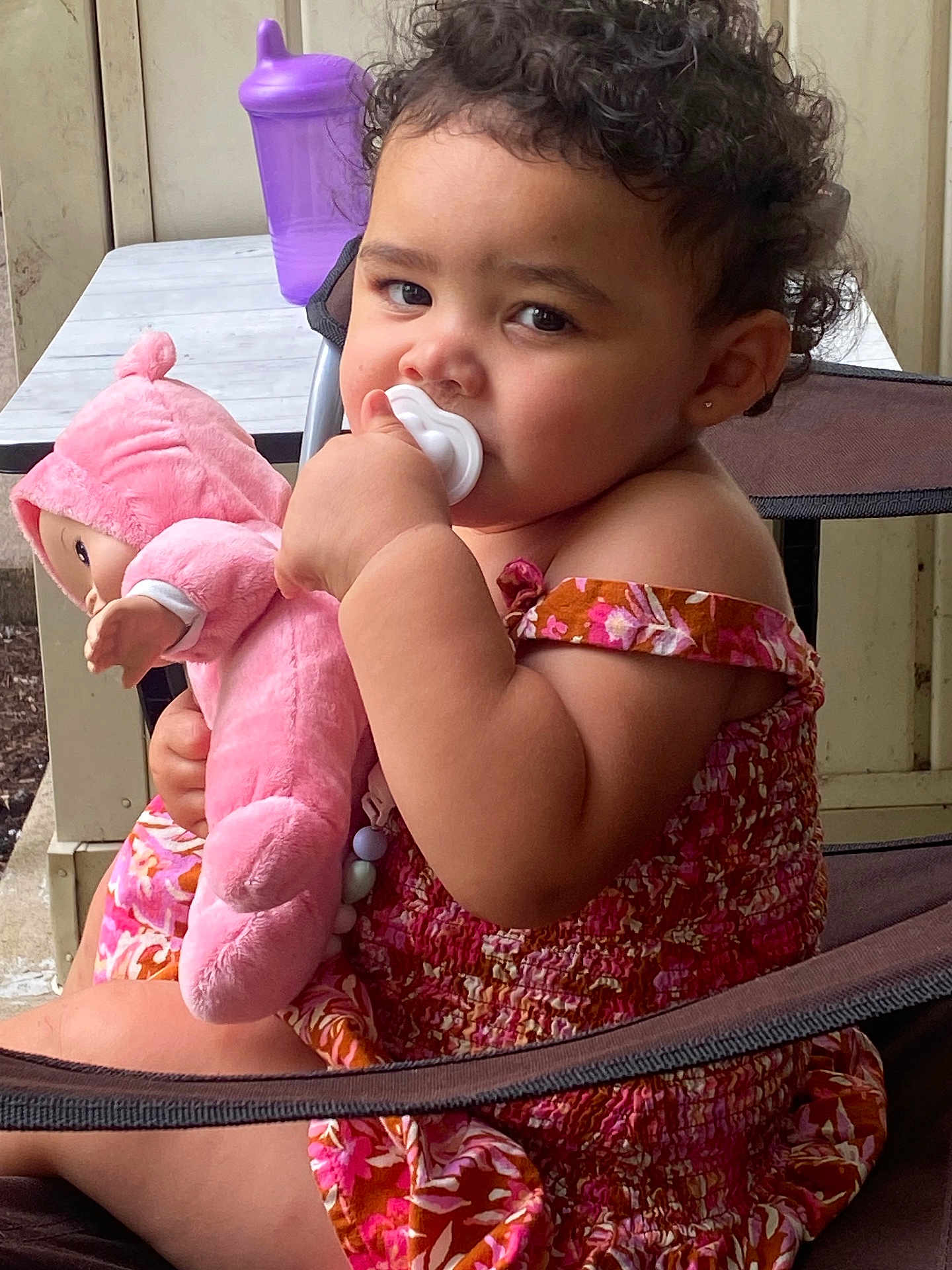 Nevayla is registered to the contest to win money with this photo: toddler, child, pacifier, plush_doll, curly_hair, pink_clothing, floral_pattern, chair, sippy_cup, table, porch, outdoor, baby, cute, person, portrait, sitting, young_child, indoor_outdoor, furniture
