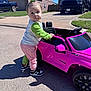 Chloe is registered to the contest to win money with this photo: casual_clothing, child, curious, daytime, driveway, grass, green_hair_bow, house, outdoor, pink, play, shadow, sidewalk, sneakers, standing, suburban, sunny, toddler, toy_car, vehicle