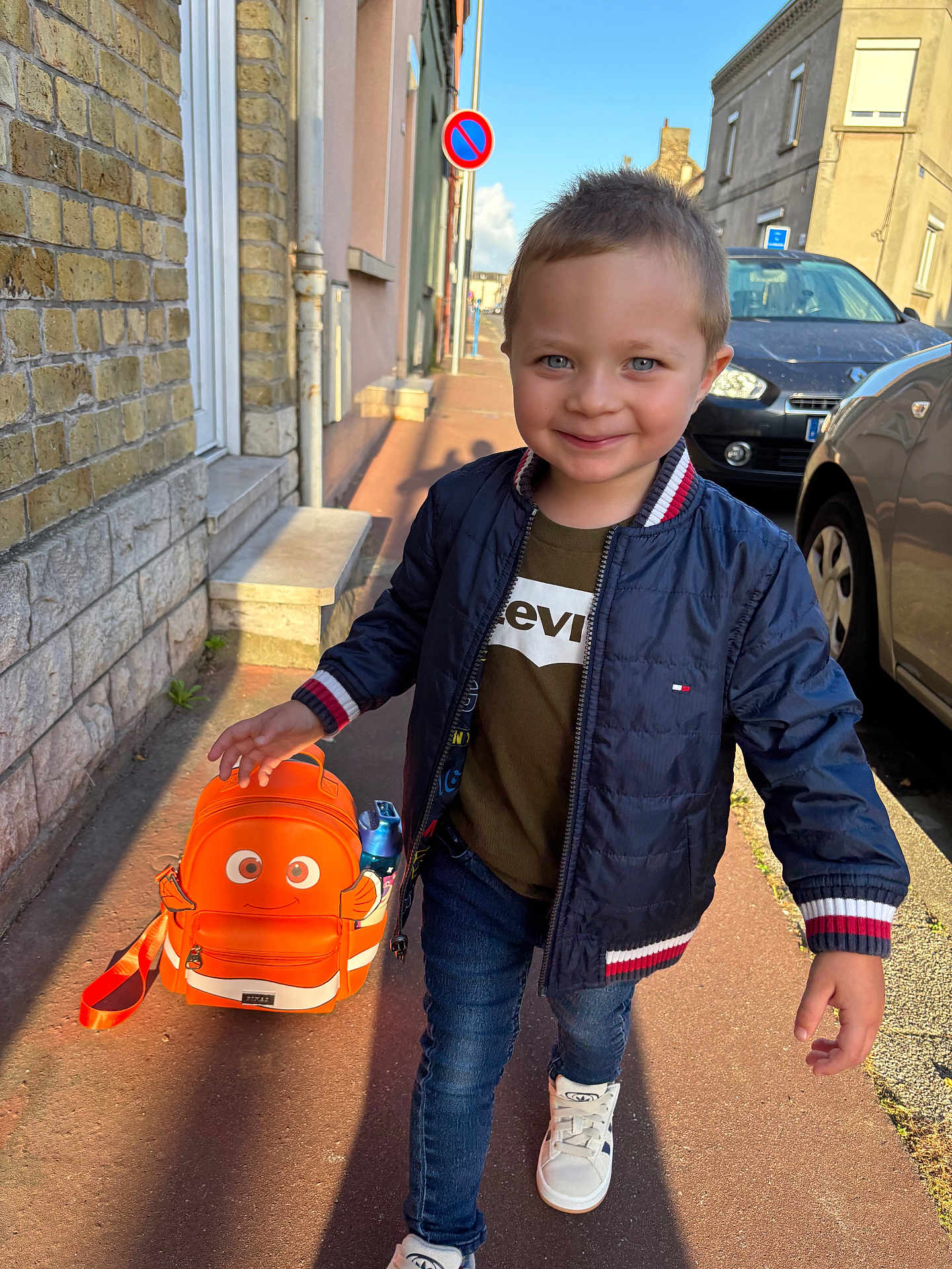 Ethan a rejoint le concours — aidez-le/la à gagner de superbes lots ! child, boy, backpack, orange, street, sidewalk, smiling, blue_jacket, jeans, sneakers, daylight, urban, car, building, window, happy, walking, person, sunlight, casual_clothing
