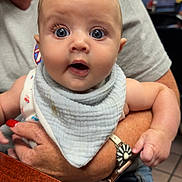 Karson is registered to the contest to win money with this photo: baby, infant, child, person, wide_eyes, surprised_expression, bib, clothing, adult_arm, watch, hand, nail_polish, indoor, table, blurred_background, skin, face, cute, holding, portrait