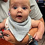 Karson is registered to the contest to win money with this photo: baby, infant, child, person, wide_eyes, surprised_expression, bib, clothing, adult_arm, watch, hand, nail_polish, indoor, table, blurred_background, skin, face, cute, holding, portrait