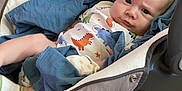 Damian is registered to the contest to win money with this photo: baby, infant, blanket, baby_seat, floor, wood_floor, clothing, onesie, dinosaur_pattern, sock, expression, face, person, indoors, cushion, fabric, cute, child, newborn, resting