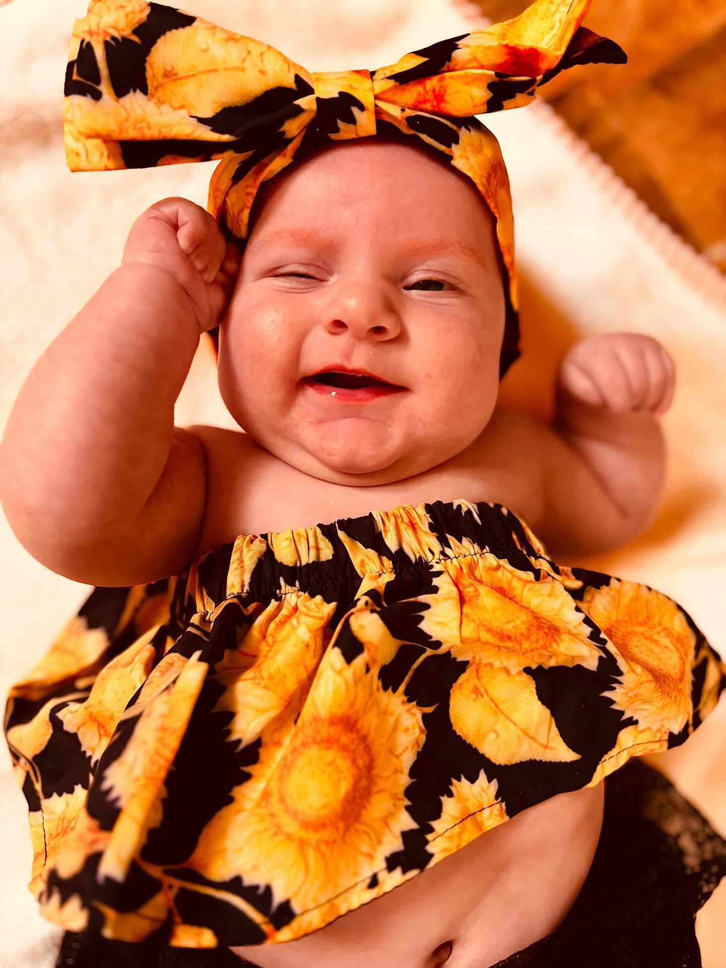 Alanni is registered to the contest to win money with this photo: baby, child, smile, winking, headband, sunflower_pattern, clothing, arm, fist, skin, face, cheeks, happy, lying_down, blanket, soft_texture, closeup, portrait, cute, infant