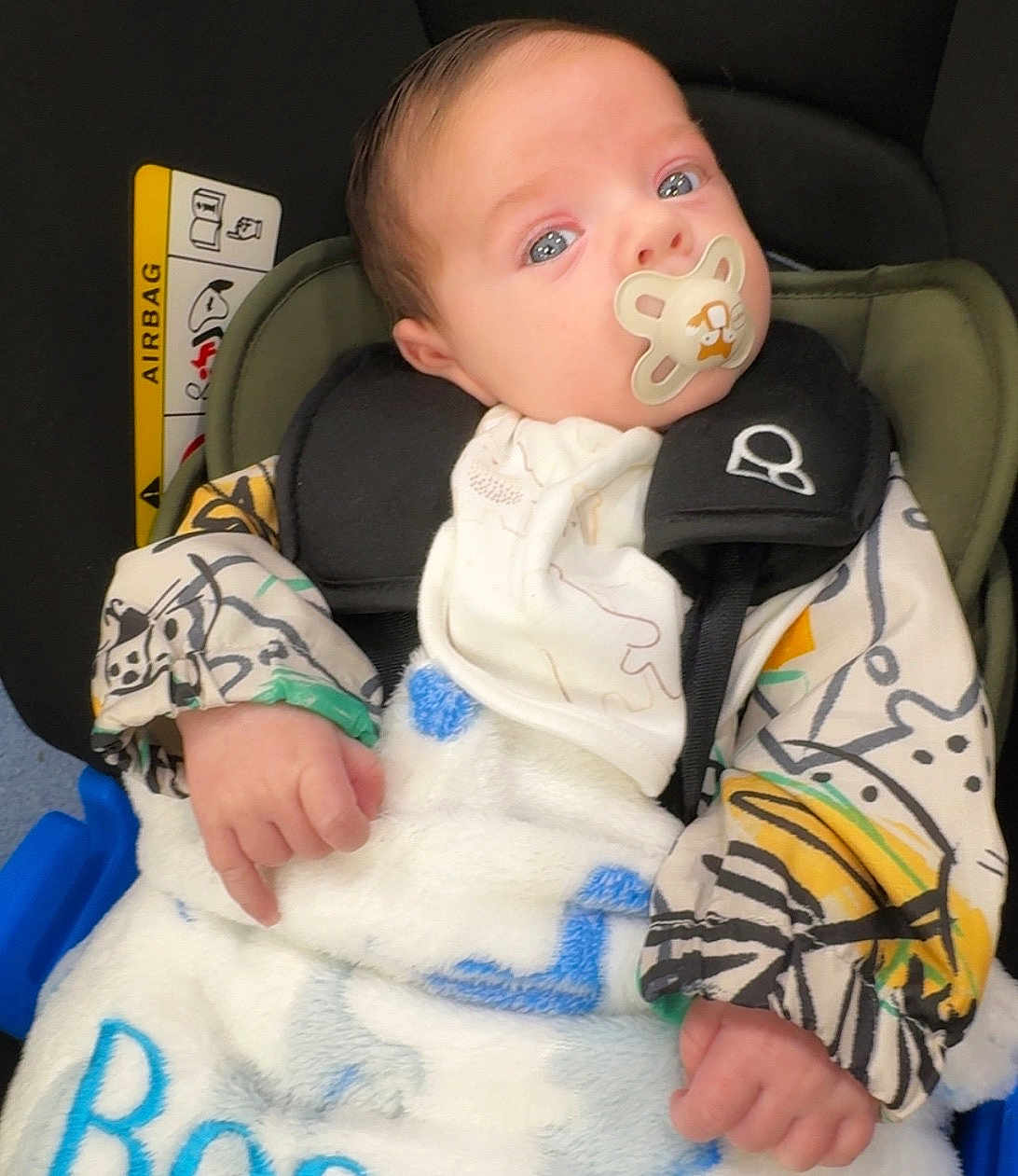 Bear is registered to the contest to win money with this photo: baby, infant, pacifier, car_seat, blanket, clothing, patterned_jacket, face, hand, child, seatbelt, safety, indoors, cute, newborn, portrait, expression, person, baby_bib, closeup