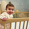 Remi is registered to the contest to win money with this photo: baby, crib, wooden_bars, smiling, child, indoor, nursery, wall_mural, hot_air_balloon, stars, cozy, cute, person, sweater, pink_pants, footwear, hair_tie, happy, portrait, standing
