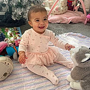 Remi is registered to the contest to win money with this photo: baby, smiling, pink_outfit, tutu, stuffed_animal, plush_toy, knitted_blanket, indoor, christmas_tree, holiday_decor, baby_play_gym, carpet, child, happy, toy, floor, infant, cute, person, playtime