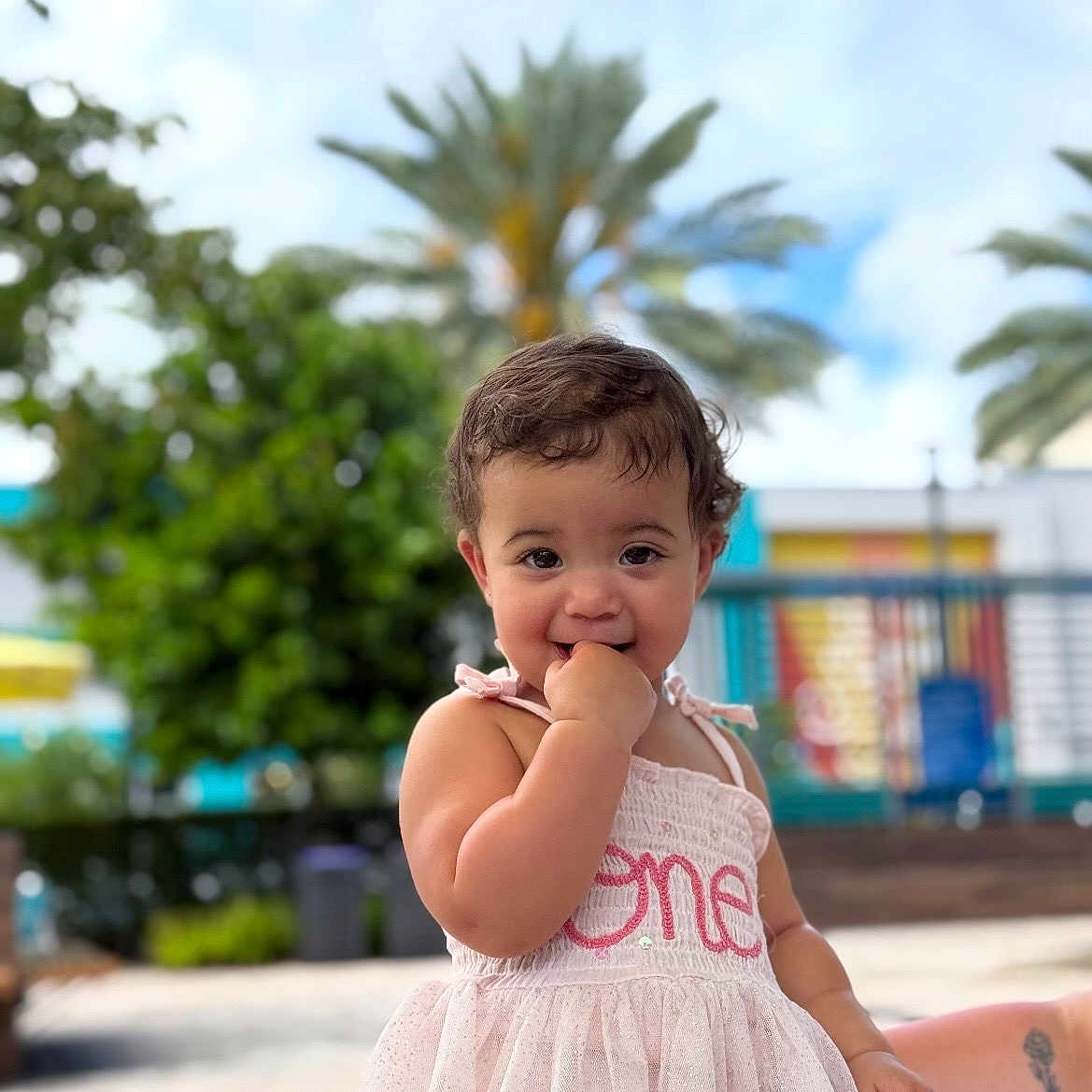 Remi joined the competition — help win amazing prizes! toddler, child, baby, dress, pink, outdoor, sunny, palm_tree, curly_hair, cute, smiling, finger_in_mouth, portrait, garden, daylight, one_year_old, playful, person, background_blur, summer
