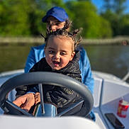 Sanad participe au concours pour gagner de l'argent avec cette photo : child, toddler, boat, steering_wheel, adult, outdoor, river, trees, blue_sky, sunny, smiling, hair_style, pigtails, clothing, watercraft, fun, family, guidance, nature, daytime