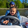 child, toddler, boat, steering_wheel, adult, outdoor, river, trees, blue_sky, sunny, smiling, hair_style, pigtails, clothing, watercraft, fun, family, guidance, nature, daytime