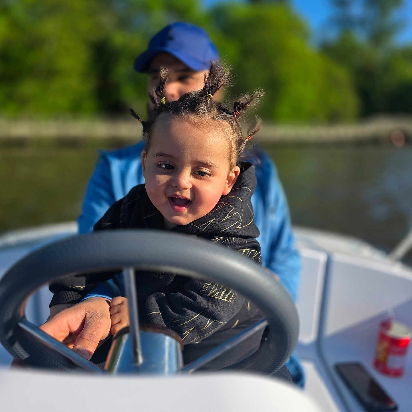 Sanad participe au concours pour gagner de l'argent avec cette photo : child, toddler, boat, steering_wheel, adult, outdoor, river, trees, blue_sky, sunny, smiling, hair_style, pigtails, clothing, watercraft, fun, family, guidance, nature, daytime
