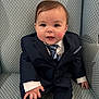 baby, child, suit, tie, armchair, smile, cute, formal_clothing, person, indoor, portrait, young_child, fashion, seated, hands, face, hair, buttoned_jacket, patterned_fabric, candid