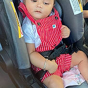 Jahnaav is registered to the contest to win money with this photo: baby, child, car_seat, red_overalls, white_shirt, bracelet, ankle_bracelet, safety_belt, seat_belt, car, infant, cute, portrait, indoors, seat, young_child, person, clothing, hand, foot