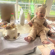 Farel a rejoint le concours — aidez-le/la à gagner de superbes lots ! baby, birthday_cake, cake, table, window, indoor, knitted_clothing, crown, toy, smiling, child, party, present, greenery, glass_bottle, cup, floor, balloon, gift_wrap, celebration